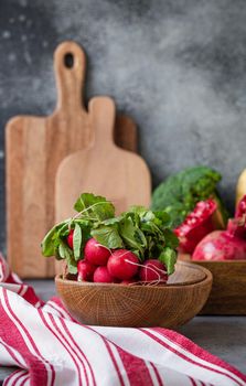 Bunch of fresh raw radish in wooden bowl on kitchen table with fresh fruit, greens, vegetables in wooden tray on grey stone background table, cooking healthy diet vegetarian food meal concept.