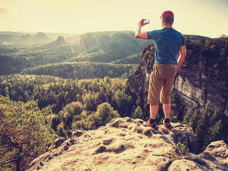 Hipster hiker. Amateur takes photos with smart phone on peak of rock. Dreamy fogy landscape spring sunrise 