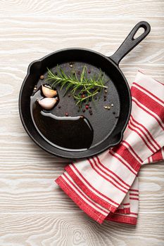 Black cast iron frying pan with ingredients for cooking: oil, fresh green rosemary, raw garlic cloves and colorful peppercorns on white rustic wooden background flat lay top view