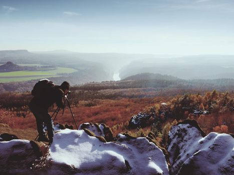 Professional photographer takes photos with mirror camera and tripod on snowy peak. Dreamy fogy landscape, spring orange pink misty sunrise in beautiful valley below.