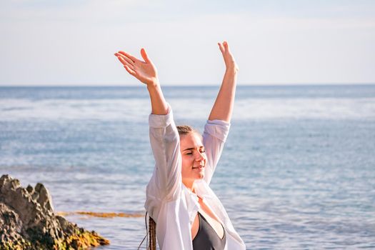 The girl stands on the shore and looks at the sea. Her hands are raised up. She wears a white shirt and her hair is in a braid