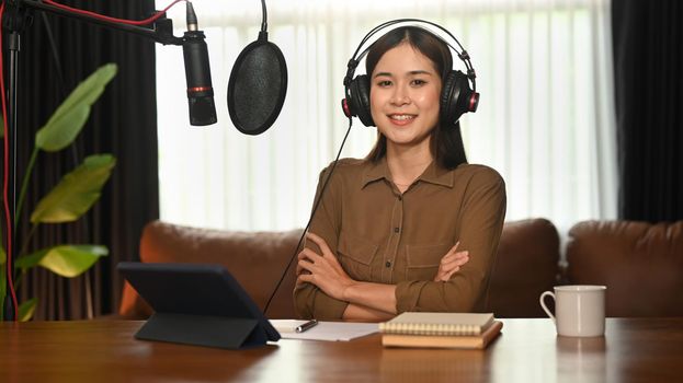 Confident female podcaster wearing headphone sitting front of condenser microphone and laptop at her studio.