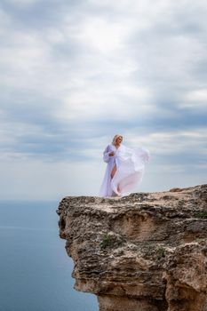 Blonde with long hair on a sunny seashore in a white flowing dress, rear view, silk fabric waving in the wind. Against the backdrop of the blue sky and mountains on the seashore