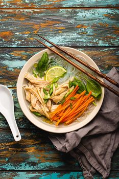 Asian soup with rice noodles, chicken and vegetables in ceramic bowl served with spoon and chopsticks on rustic wooden background from above, Chinese or Thai cuisine