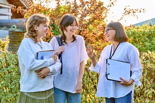 Female teacher talking to teenage female students outdoor. Two girls 17, 18 years old with middle age mentor. Teaching, education, youth, high school, college, university concept