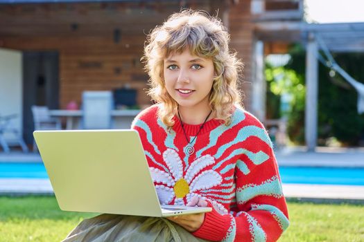 Portrait of teenage girl with laptop outdoor, smiling cute female teenager posing looking at camera, sitting on backyard lawn. Beautiful girl 16, 17 years old with curly hair, high school student