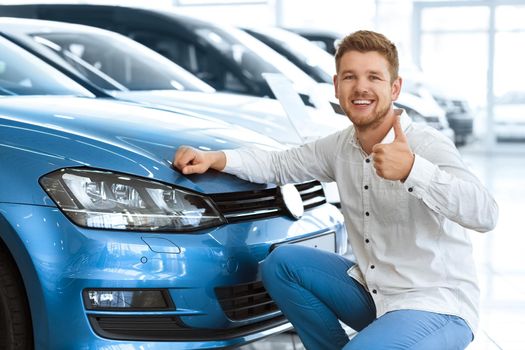 Plus one driver on the road! Happy young man showing thumbs up posing near his newly bought car at the dealership