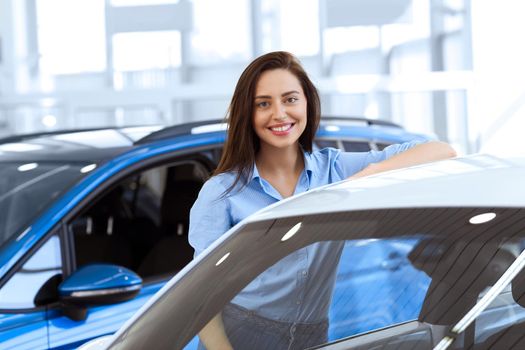 Perfect car for me. Gorgeous young woman smiling happily leaning on a car at the local car dealership
