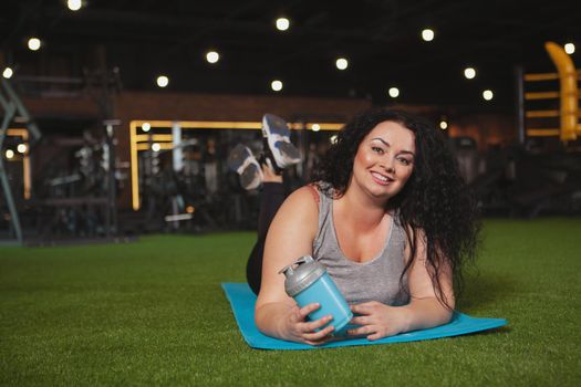 Cheerful plus size woman relaxing at the gym after working out, lying on yoga mat, copy space. Happy beautiful sportswoman resting at sport studio