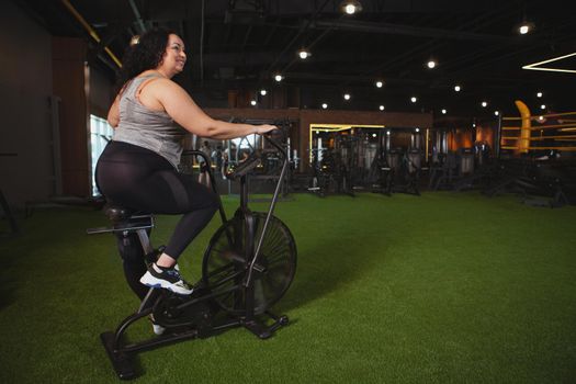Rear view shot of a plus size sportswoman smiling, looking away, cycling on air bike at the gym, copy space. Cheerful overweight woman enjoying cardio workout at sport studio