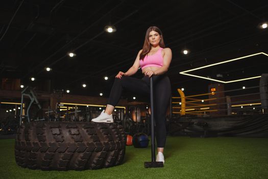 Young attractive plus size woman looking confidently to the camera, resting after working out with sledgehammer and heavy huge wheel tire. Gym, body positive concept