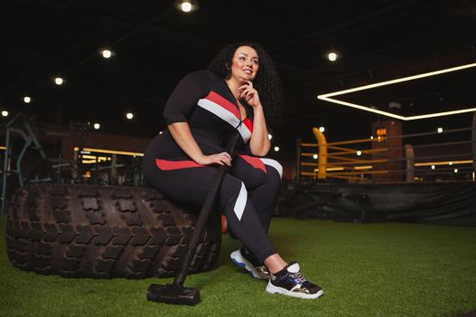Low angle shot of a plus size woman in sportswear resting at the gym after exercising with sledgehammer. CHeerful overweight sportswoman relaxing after hitting heavy wheel with a hammer, copy space