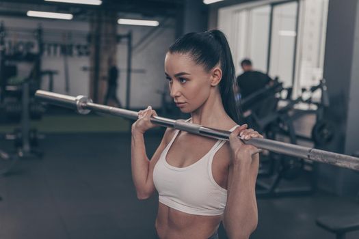 Gorgeous fit and toned sportswoman lifting barbell at the gym, copy space on the side. Attractive female athlete in white sports bra exercising with barbell. Sexy sportswoman weightlifting
