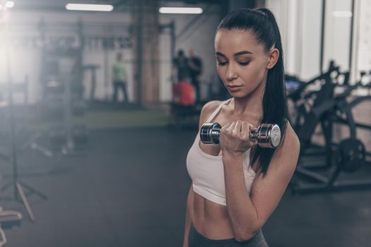 Young attractive fitness woman looking concentrated, exercising with dumbbells at the gym. Sexy sportswoman lifting weights at sports studio, copy space