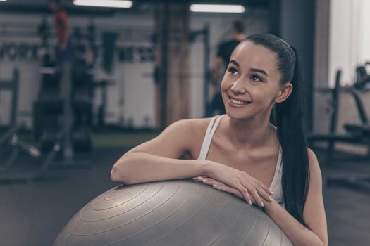 Charming happy young woman resting at the gym after exercising on fitness ball, smiling looking away joyfully. Cheerful beautiful sportswoman relaxing after workout, copy space. Health concept