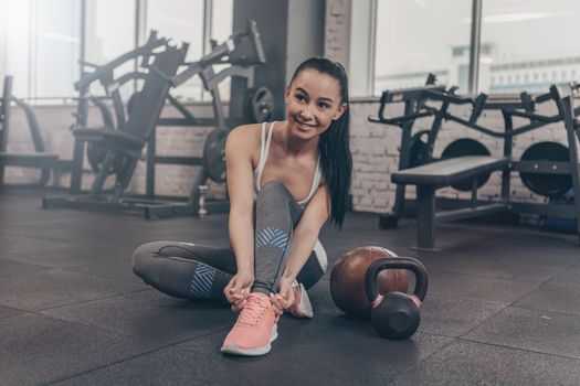 Full length portrait of a beautiful happy healthy woman tying her shoelaces, sitting on the gym floor. CHeerful fitness female resting after workout, tying her sneakers, copy space