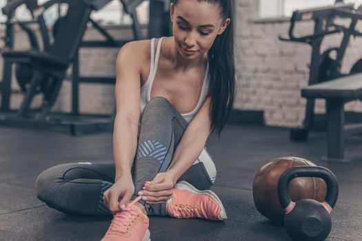 Cropped shot of a lovely young sportswoman tying her shoelaces, sitting on the gym floor, copy space. Beautiful fitness woman resting after working out at sports studio. Motivation, sportswear concept