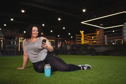 Lovely plus size sportswoman using her smart phone at the gym, copy space. Cheerful curvy woman in sportswear resting after exercising