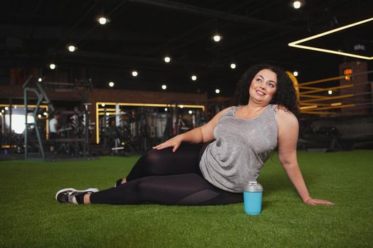 Beautiful healthy happy plus size woman sitting on the floor at the gym, resting after exercising, copy space. Lovely curvy fitness woman relaxing after intense workout