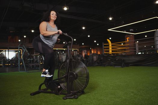 Body positive, feminine confidence concept. Full length shot of a overweight beautiful happy woman cycling at the gym on air bike. Cheerful plus size woman doing cardio workout on air cycle, copy space