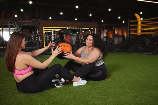 Cheerful overweight women in sportswear passing medicine ball to each other, doing abs crunches, copy space. Happy plus size women enjoying exercising at sports studio together