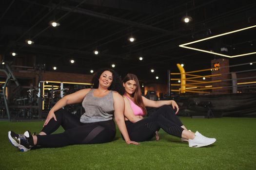 Two beautiful healthy happy plus size fitness women resting at the gym after exercising, copy space. female friends relaxing after workout