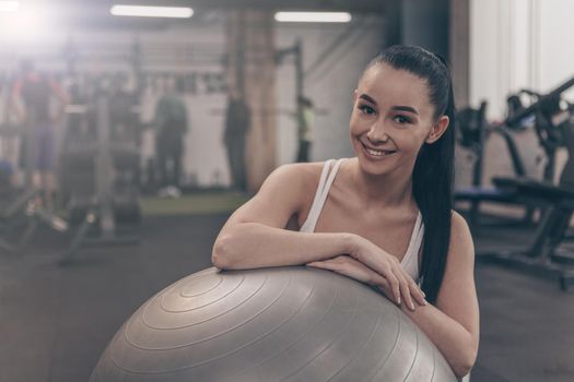 Young happy beautiful woman smiling to the camera, resting at the gym after working out with fitness ball. Cheerful healthy sportswoman relaxing at sports studio after exercising, copy space