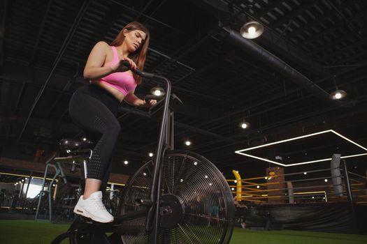 Low angle shot of a plus size woman in sportswear cycling on air bike. Young sportswoman doing cardio workout on air bicycle at the gym, copy space