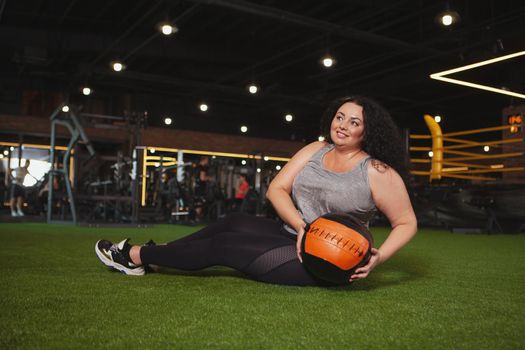 Happy mature plus size woman exercising with medicine ball at the gym. Cheerful beautiful overweight woman doing abs exercise at sports studio