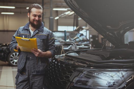 Friendly bearded male mechanic enjoying working at his garage, taking notes on clipboard, looking under the hood of a car. Cheerful auto technician repairing cars at his workshop, copy space