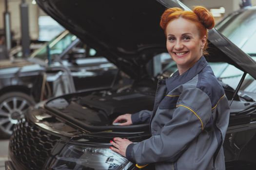 Lovely female mechanic smiling to the camera over her shoulder, while examining broken car with an open hood. Beautiful happy red haired woman working at car service station, repairing cars, copy space. Occupation, hobby, vehicle concept
