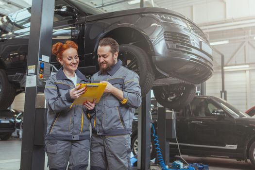 Cheerful female mechanic talking to her male colleague, working at car service station, copy space. Bearded auto technician discussing work with his female colleague, car on a lift on background