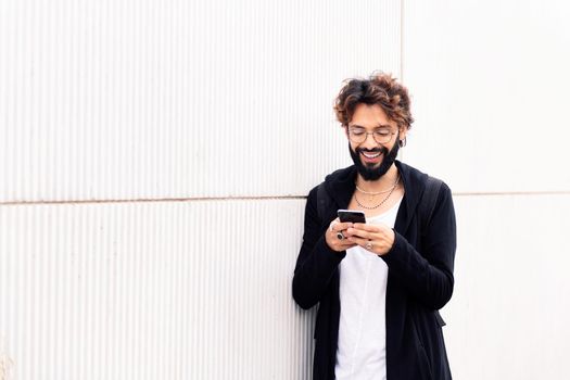 smiling young man with beard leaning in a white wall typing happy on mobile phone, concept of urban lifestyle and technology of communication, copy space for text