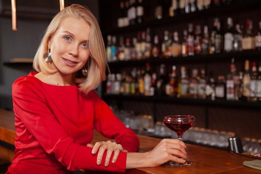 Beautiful elegant woman in red dress smiling to the camera, enjoying her cocktail at local bar