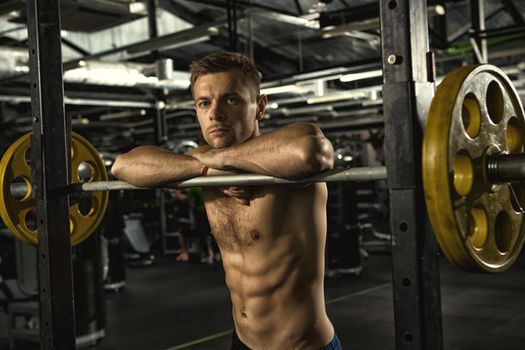 Resting in between sets. Young handsome shirtless sportsman looking to the camera leaning on the barbell resting after his workout at the gym exercising training fitness healthcare confidence concept