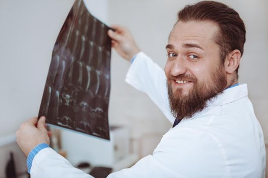 Close up of a cheerful bearded male doctor smiling to the camera over his shoulder, while checking mri scans of a patient. Happy practitiner examining x-ray scans of a patient