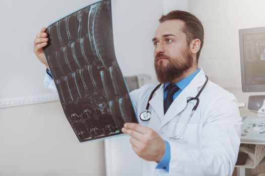 Attractive bearded male doctor concentrating, examining mri scans of a patient. Experienced therapist checking results of an x-ray of a patient. Health, medicine, treatment concept