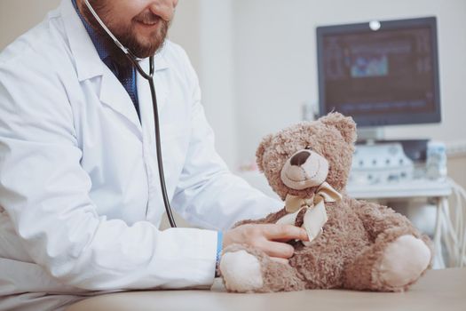 Cropped shot of unrecognizable male doctor smiling, examining plush toy teddy bear, using stethoschope, copy space. Pediatrician working at the clinic. CHildhood, motherhood, parenthood concept