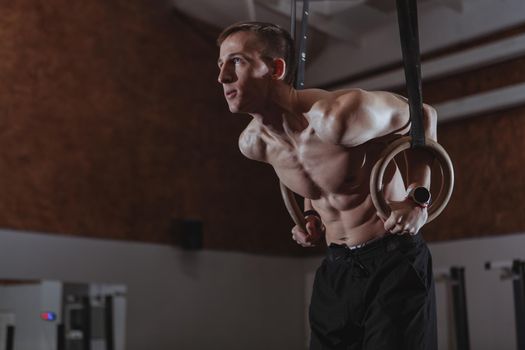 Shirtless male crossfit athlete performing ring dips at the gym. Athletic man looking concentrated, working out on gymnastic rings, copy space