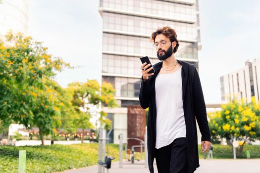 young man with beard walking by the city using a mobile phone, concept of urban lifestyle and technology of communication, copy space for text