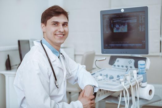 Charming young male doctor smiling to the camera, sitting at his office near ultrasound scanning machine. Handsome friendly doctor enjoying working at the hospital, copy space. Ultrasonic concept