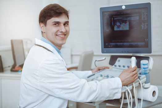 Happy handsome male doctor smiling to the camera, working on ultrasound scanning machine at his clinic. Experienced doctor working at modern medical facility with high quality equipment