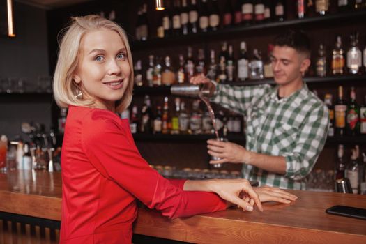 Beautiful happy blond woman smiling to the camera, waiting for her cocktail at the bar