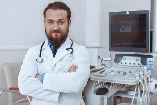 Confident male doctor posing proudly at his clinic near ultrasound scanner, copy space. Successful experienced doctor at his office. Technology, modern medicine, health care concept