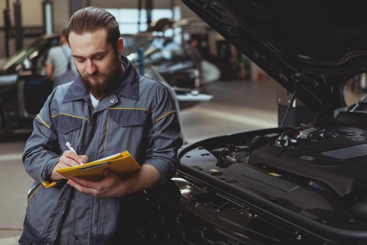 Bearded male mechanic looking concentrated, writing on his clipboard, leaning on a car with an open hood. Experienced car service worker filling papers after repairing automobile, copy space