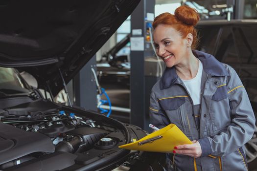 Beautiful woman working at car repair service, taking notes on clipboard. Attractive female technician examining engine of an auto. Female mechanic looking under the hood of a car