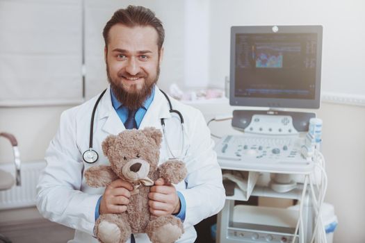 Happy male bearded doctor smiling to the camera, holding plush teddy bear toy, copy space. Cheerful pediatrician enjoying working at kids clinic. Pediatrics, childhood, health concept