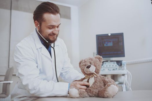 Bearded male doctor concentrating, using stethoscope, examining cute teddy bear. Friendly male pediatrician working at his clinic, pretending to examining teddy bear toy, copy space