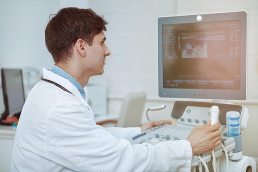 Rear view shot of a male doctor using ultrasound scanning machine at his clinic. Practitiner working on modern medical equipment at his office. Ultrasound diagnostics, pregnancy, cancer concept