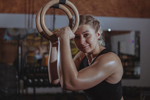 Charming female crossfit athlete smiling to the camera, holding gymnastic rings. Sportswoman smiling joyfully relaxing at crossfit gym after exercising, copy space